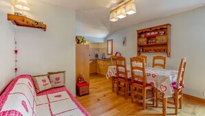 A cozy room with a bed, a dining table with chairs, a kitchen area, and a wooden shelf with decorative items.