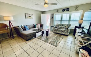 A living room with a grey sofa, a patterned armchair, a coffee table, and a floor lamp. There is a door leading outside, a window with blinds, and a ceiling fan. - Sea Sounds | ★Beachfront★ | 2 Pools | First Floor Island Retreat 2 Bedroom Condo (Port Aransas)