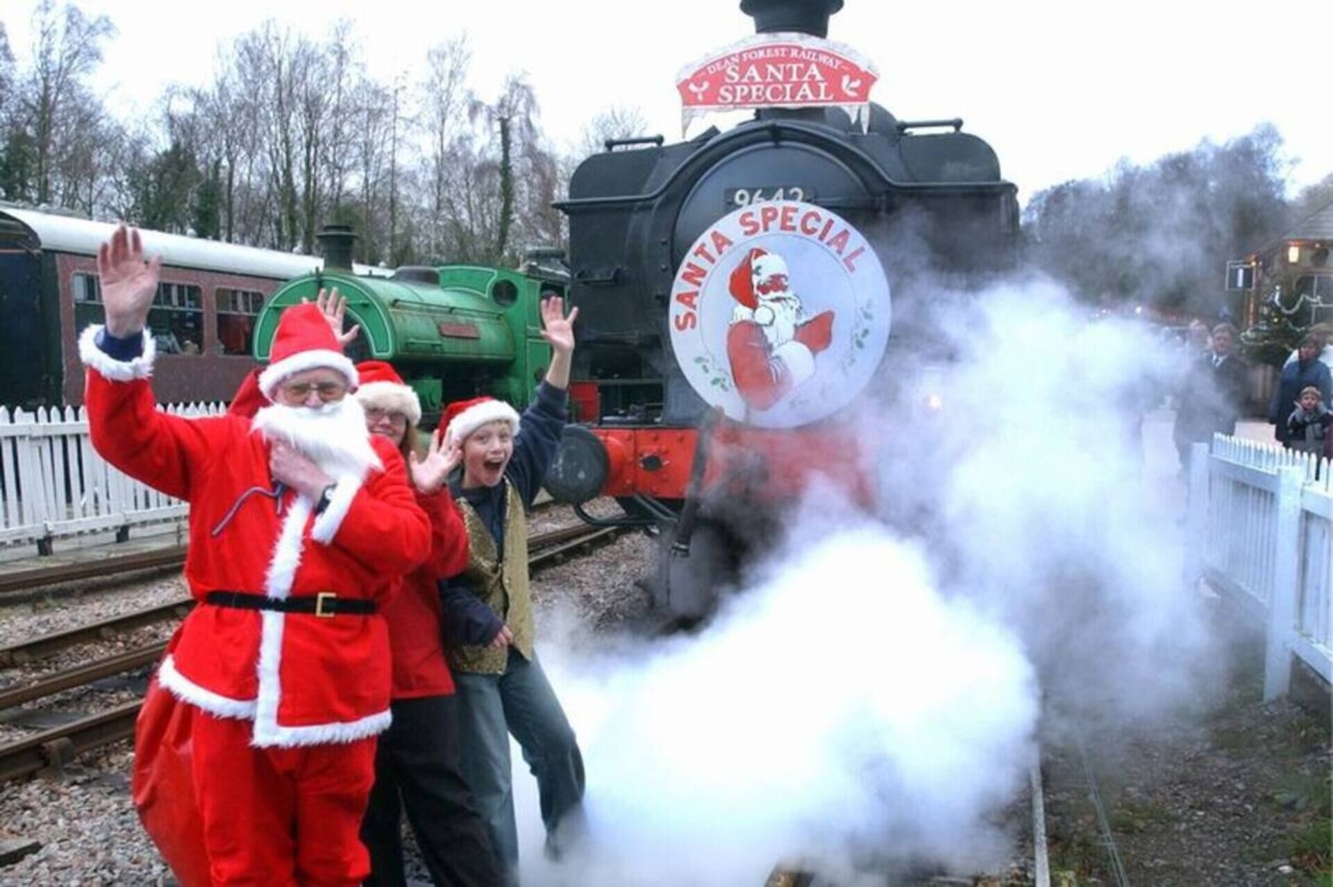 A steam locomotive with a Santa Claus logo, surrounded by people in festive attire.
