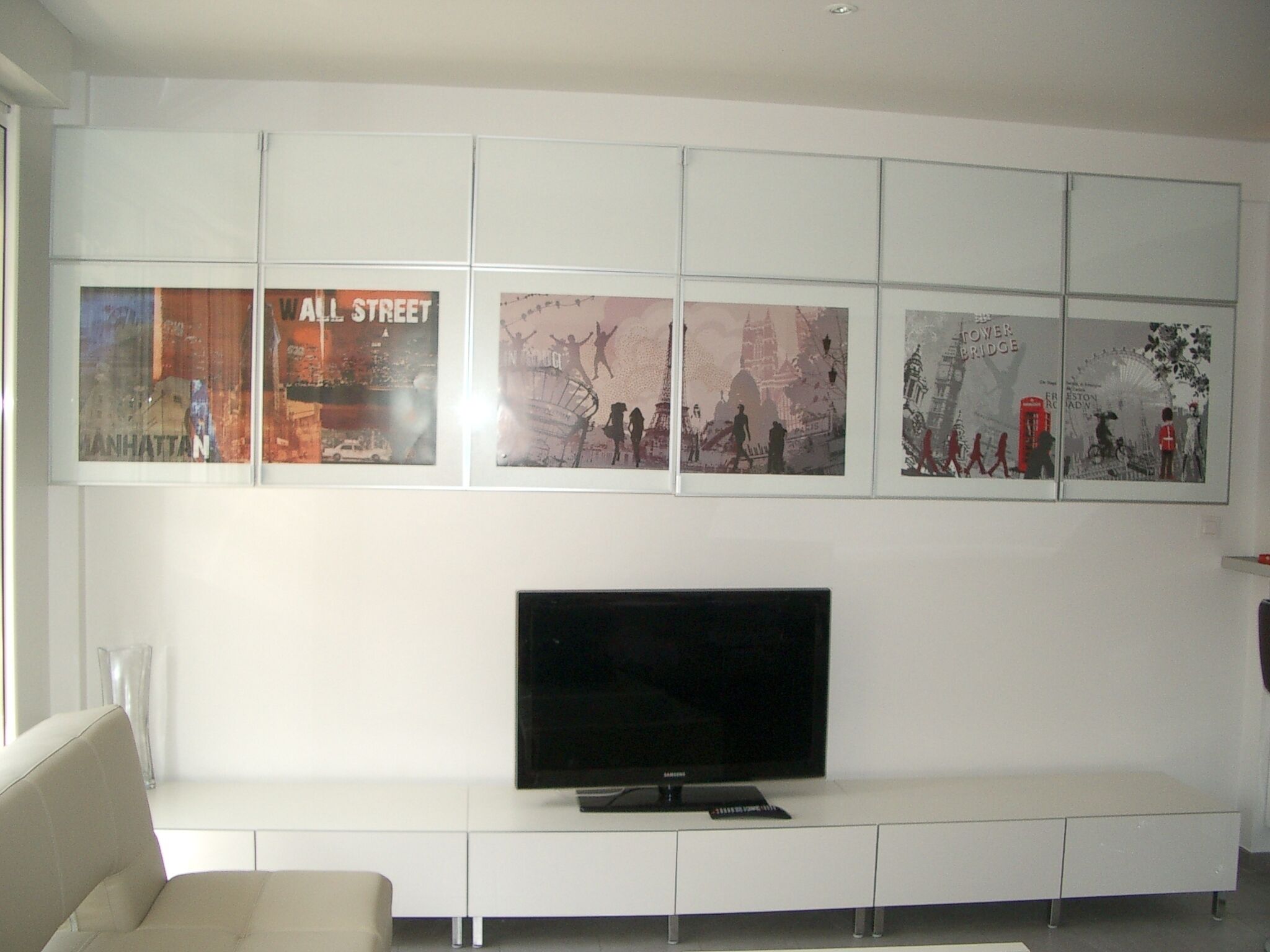 A modern living room with a flat-screen TV on a white entertainment unit, above which are framed cityscape posters.