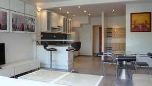 A modern kitchen with white cabinets, a microwave, and a sink. There are two bar stools and a dining table with chairs. The room has a flat-screen TV and a painting on the wall. - 2 /3 ROOMS NEAR BEACH AND CONVENTION CENTER 4 PEOPLE MAXIMUM (Alpes-Maritimes)
