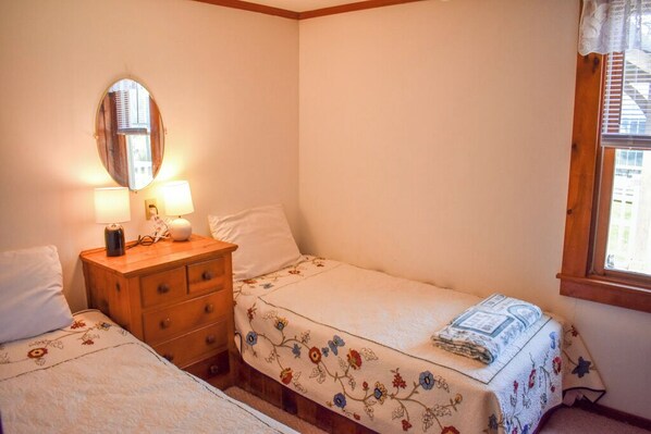 A small, cozy bedroom with two single beds, a wooden dresser, a mirror, and a window with blinds.