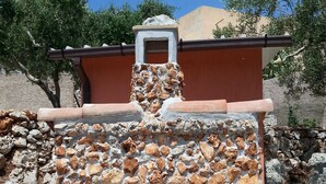 Una chimenea de piedra al aire libre con un tiro, rodeada por un muro de piedra y un edificio rojo al fondo.