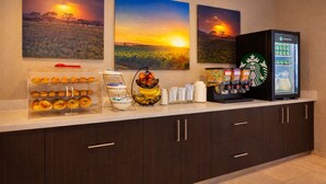 A hotel breakfast area with a coffee machine, fruit bowl, and bagel display. - Days Inn & Suites by Wyndham Tomball/Houston NW (Tomball)