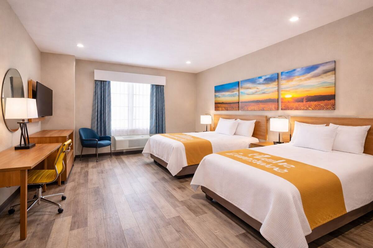 A hotel room with two beds, a desk, a chair, a TV, and a large window with curtains.