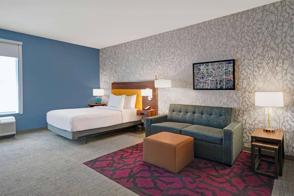 A hotel room with a bed, a sofa, an ottoman, a nightstand, a lamp, and a wall map.