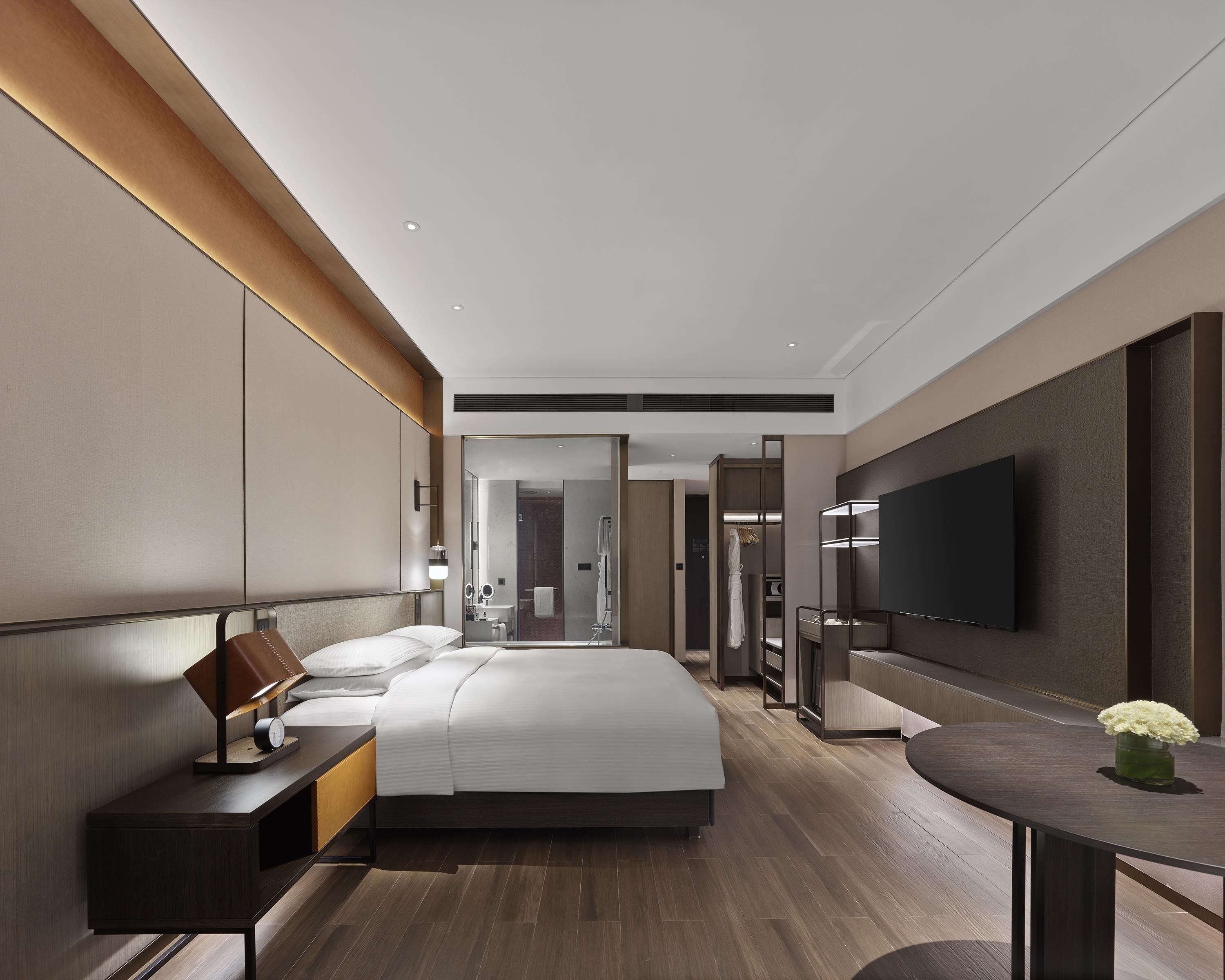 Quarto de hotel moderno com uma cama grande, mesa de cabeceira, luminária e televisão na parede.