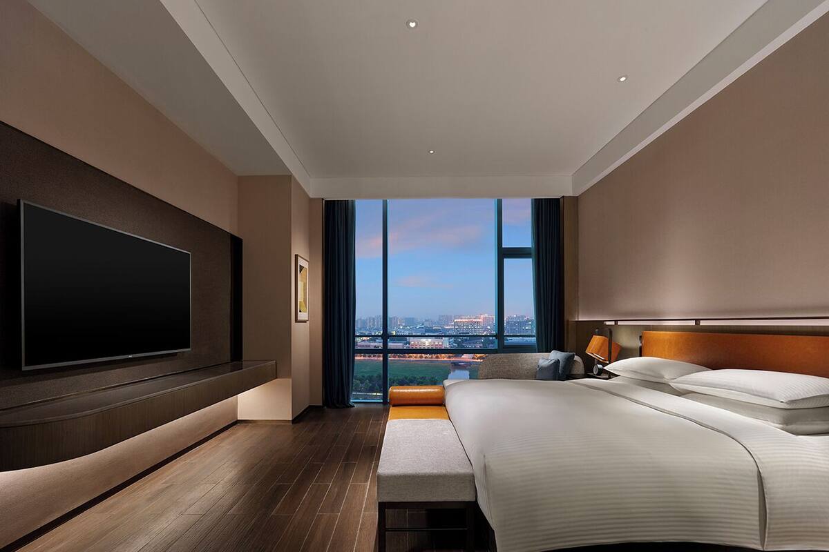A modern hotel room with a large bed, a flat-screen TV mounted on the wall, and a city view through the window.