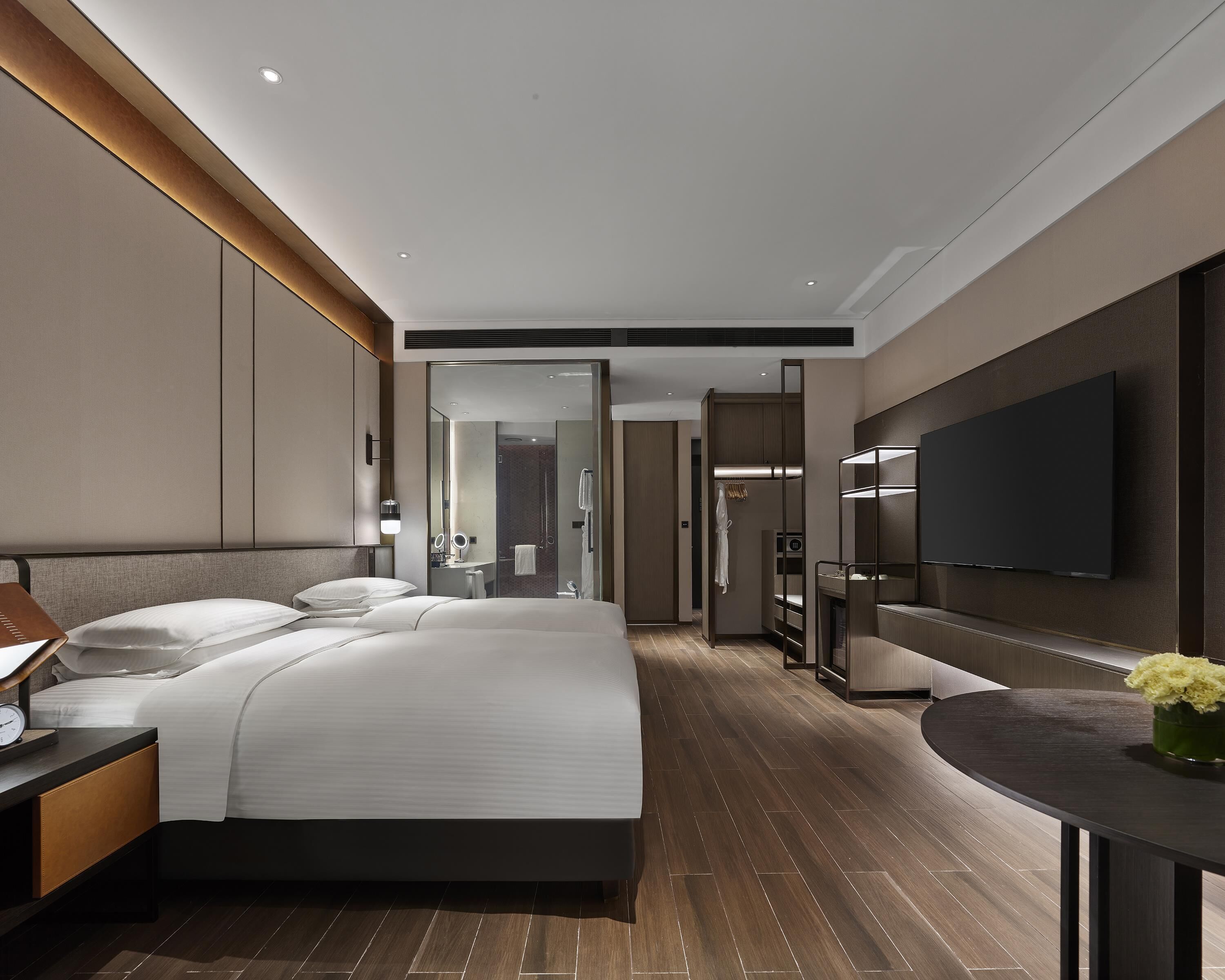 A modern hotel room with a large bed, a flat-screen TV, a desk with a lamp, and a bathroom visible through a glass door.