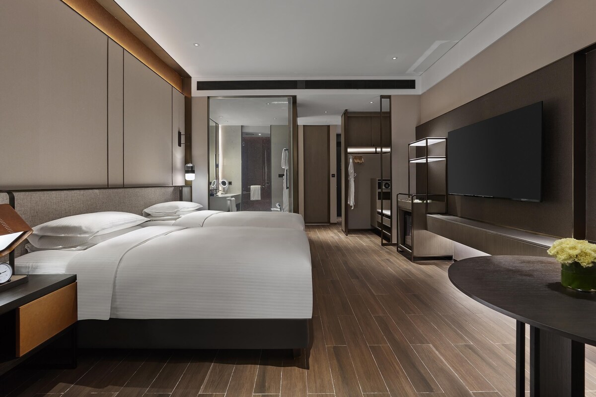 Una habitación de hotel moderna con una cama grande, un televisor de pantalla plana, un escritorio con lámpara y un baño visible a través de una puerta de cristal.