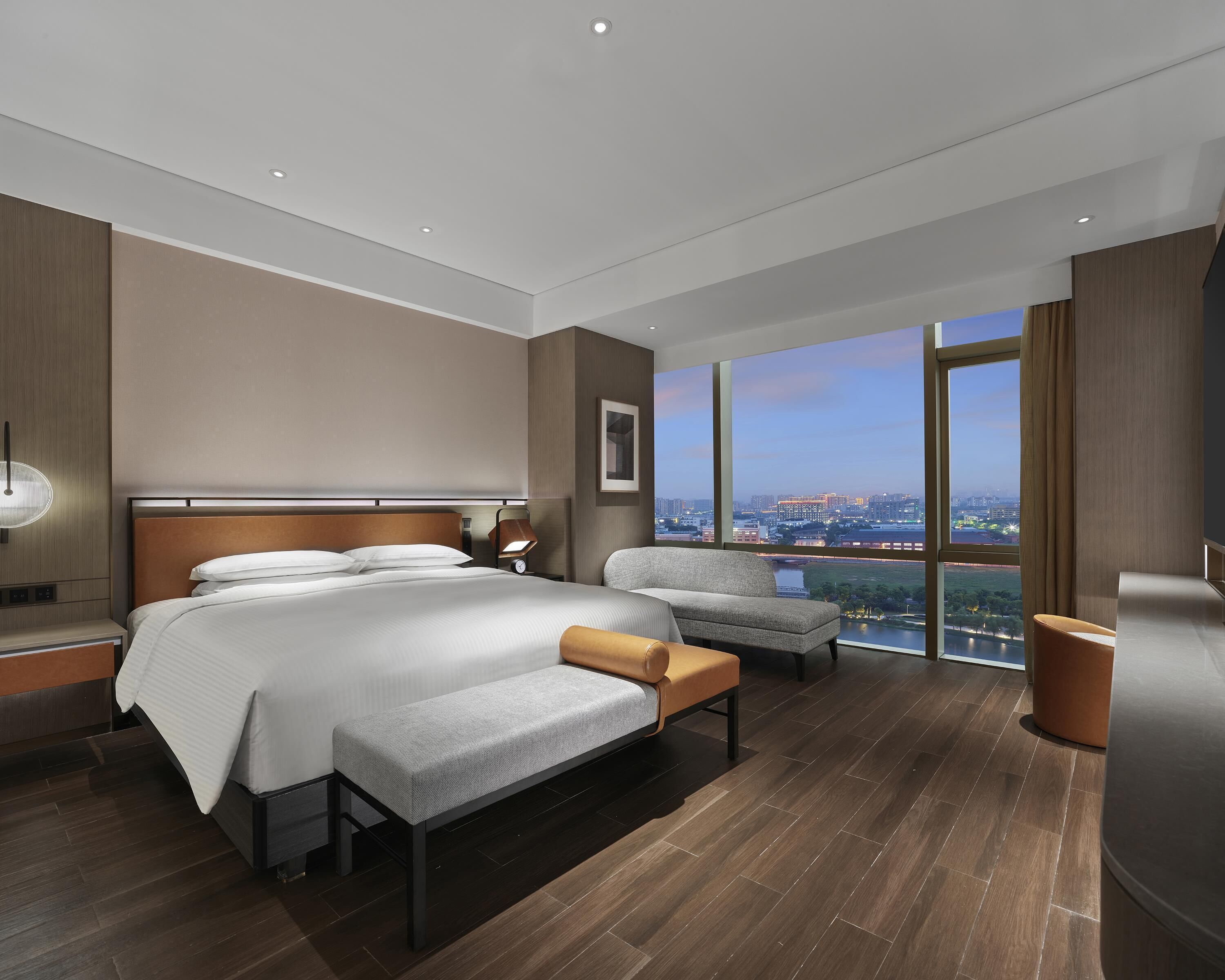 Ein modernes Hotelzimmer mit einem großen Bett, einer Sitzecke, einem Nachttisch, einer Lampe und Blick auf die Stadt.