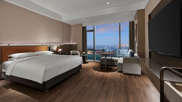 A modern hotel room with a large bed, a seating area with a view of the city, and a flat-screen TV.