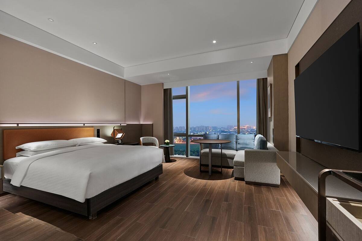 A modern hotel room with a large bed, a seating area with a view of the city, and a flat-screen TV.