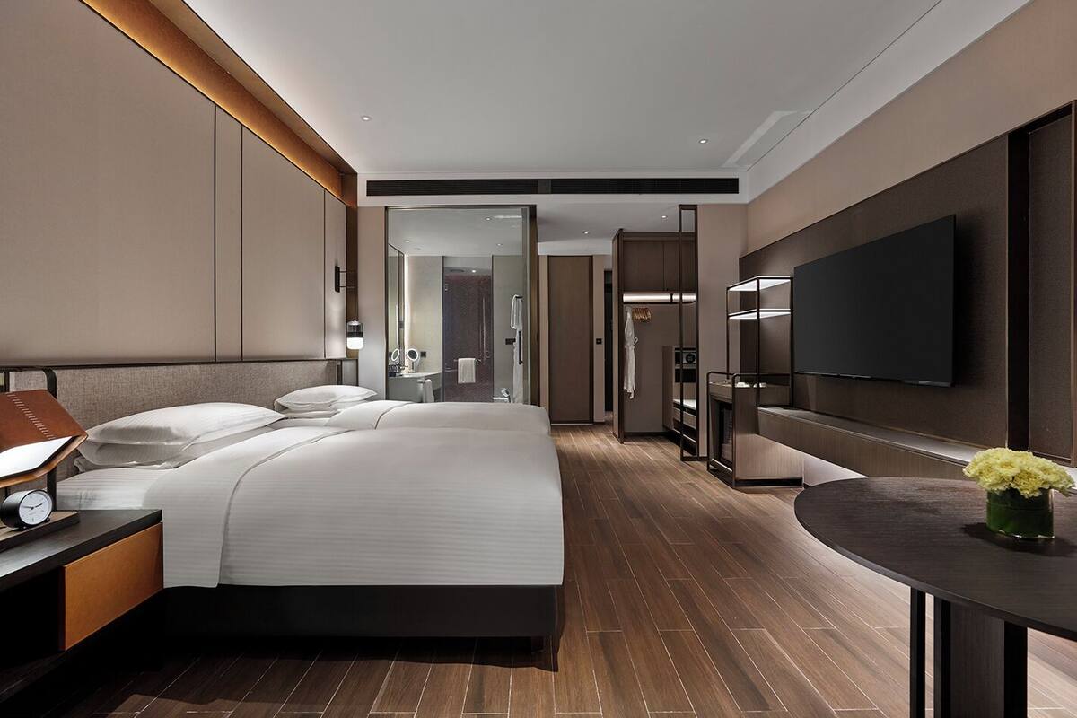 A modern hotel room with a large bed, a bedside table with a lamp, a TV mounted on the wall, and a small table with a vase of flowers.