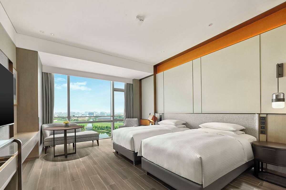 A modern hotel room with two beds, a TV, a balcony with a view of the city, and a dining area with a table and chairs.