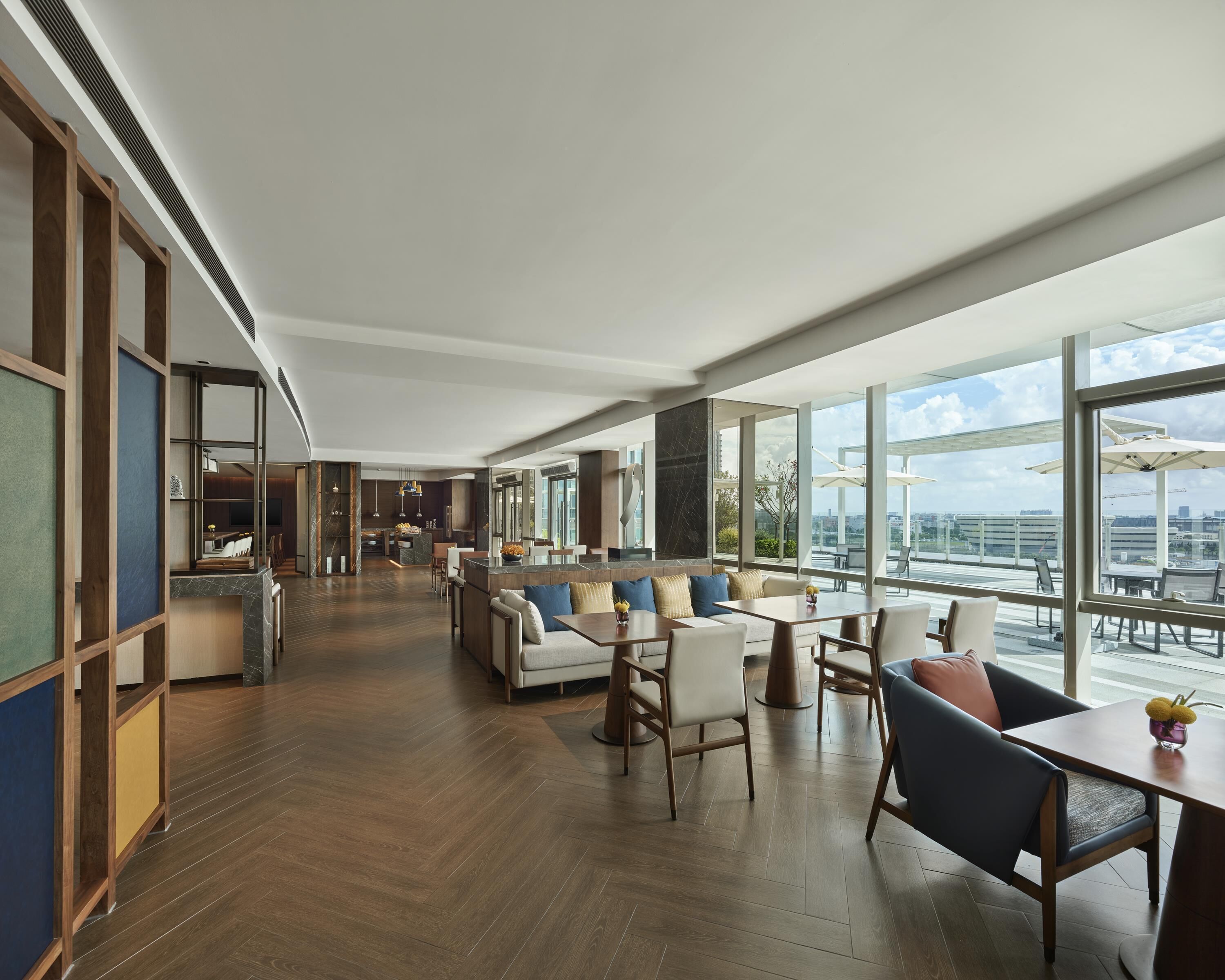 Un amplio vestíbulo de hotel con techo alto, piso de madera, zona de estar con sofás y sillones, y grandes ventanas con vistas a la ciudad.