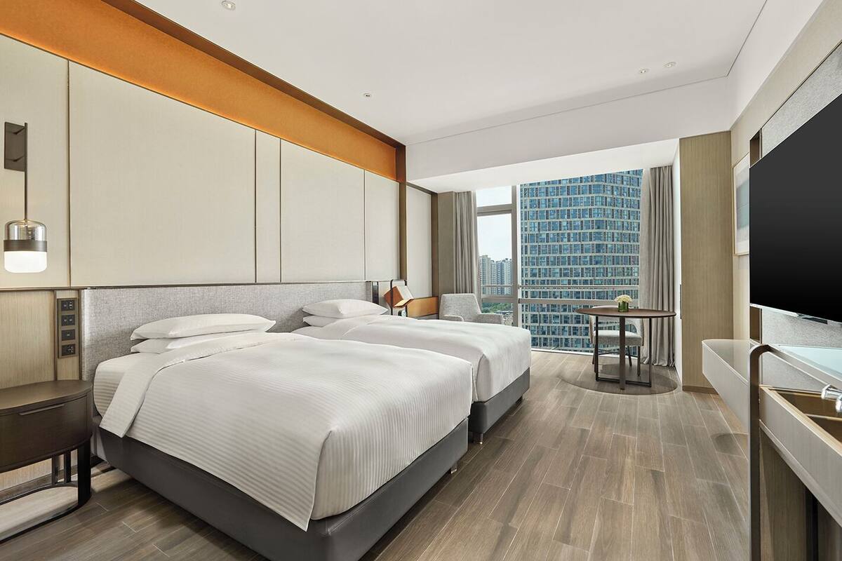 A modern hotel room with two beds, a desk, a chair, a small table with a vase, and a large window offering a city view.