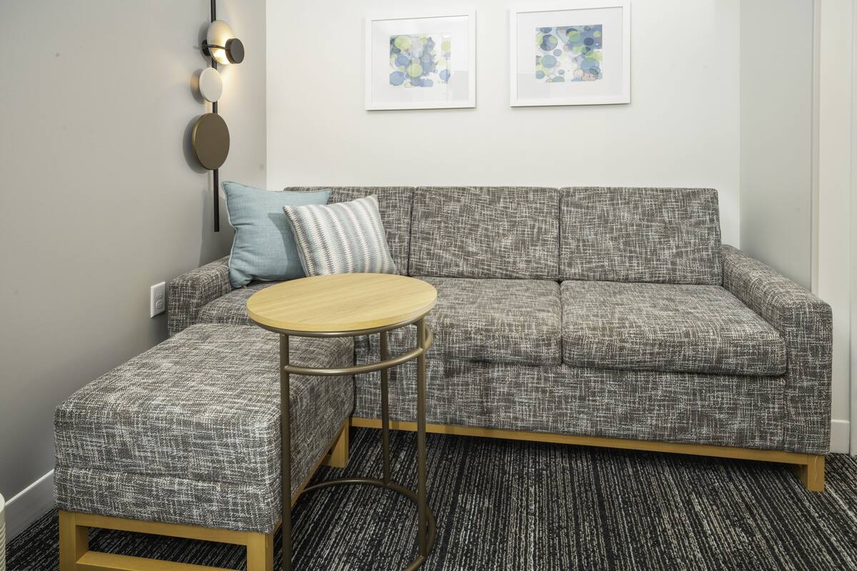 Un salon moderne avec un canapé gris à texture, une table basse ronde en bois et un pouf à motifs. Deux tableaux abstraits encadrés ornent le mur, ainsi qu’un luminaire fixé au mur.