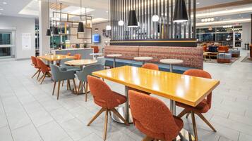 A modern dining area with wooden tables and orange chairs, a patterned sofa, and pendant lighting.