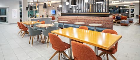 Un espace de repas moderne avec des tables en bois et des chaises orange, un canapé à motifs et des luminaires suspendus.