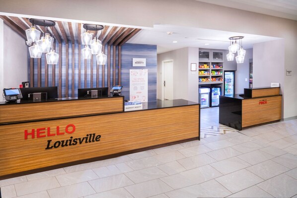 Un mostrador de recepción de hotel con un cartel que dice 'HELLO Louisville', una pantalla digital y una máquina expendedora.