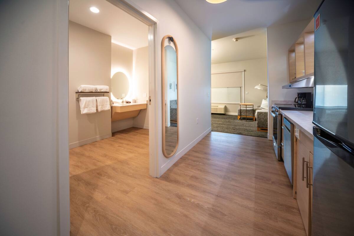 Une chambre d’hôtel moderne avec un plancher en bois, un grand miroir, une salle de bain avec un meuble-lavabo et une kitchenette équipée d’un réfrigérateur.