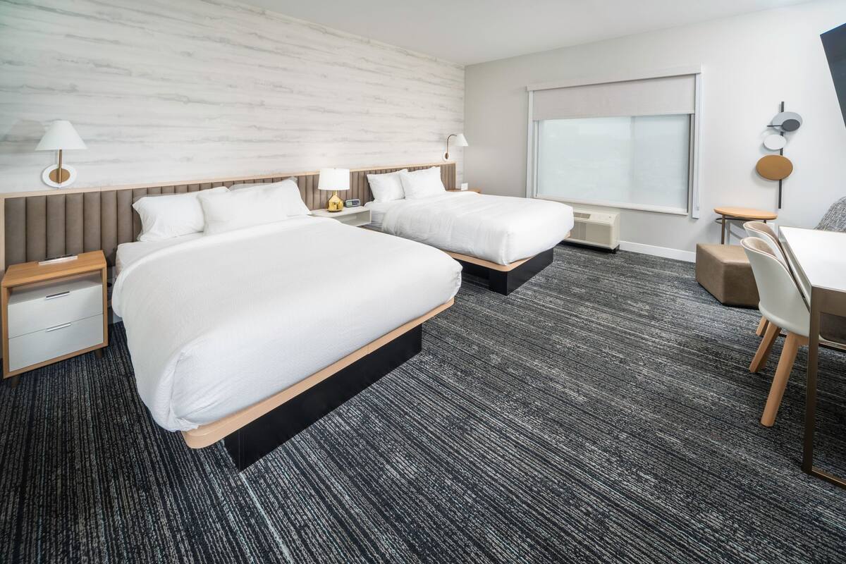 Une chambre d’hôtel avec deux lits, un bureau et une chaise. Il y a une fenêtre avec vue, une lampe et un climatiseur fixé au mur.