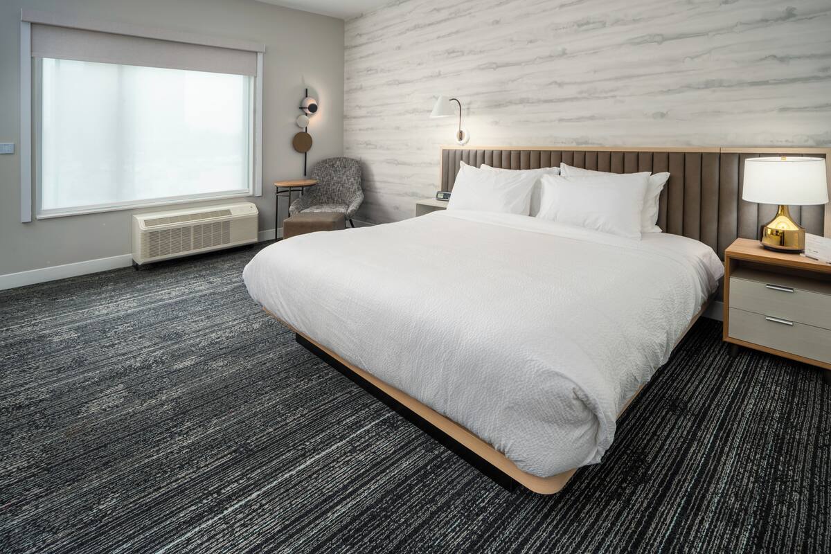 Une chambre d’hôtel moderne avec un grand lit, une chaise, une table de chevet, une lampe et une fenêtre avec des stores.