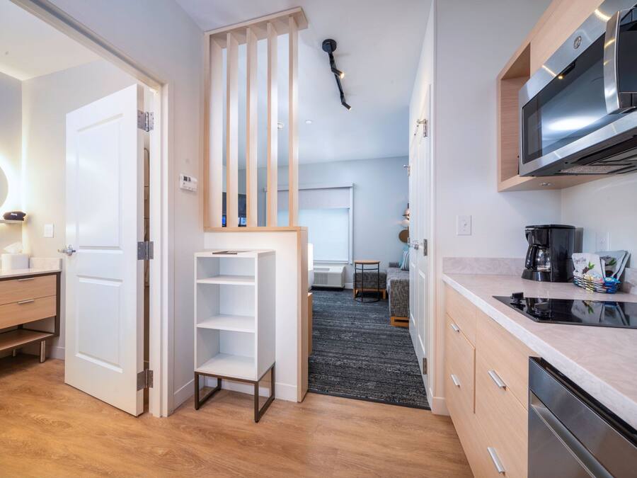 A modern kitchen with a microwave, coffee maker, and a built-in oven. There is a white door with a shelf unit and a small table in the background.