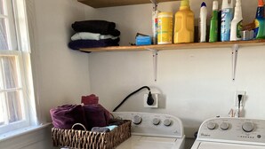 A laundry area with two white washers and dryers, a wicker basket with clothes, and shelves with cleaning supplies.