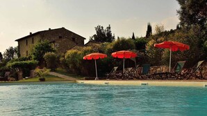 Une piscine entourée de chaises longues et de parasols rouges, le tout bordé de verdure et adossé à un bâtiment en pierre.