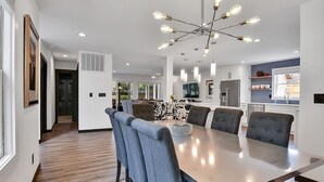 A modern dining area with a long table and six chairs, a chandelier, and a kitchen in the background.