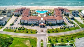 An aerial view of a beachfront hotel with a central pool, multiple buildings, and a road leading to the beach.