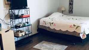 Een slaapkamer met een bed, een televisie op een standaard, een plank met diverse spullen, en een klein vloerkleed.