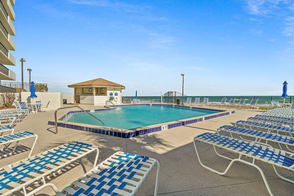 A swimming pool with lounge chairs, a small building with a clock, and a view of the ocean. - Relax & Chill Beachside Condo (Daytona Beach)