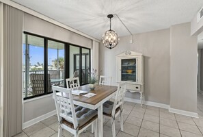 A dining area with a wooden table and white chairs, a chandelier, and a cabinet with dishes. - Relax & Chill Beachside Condo (Daytona Beach)