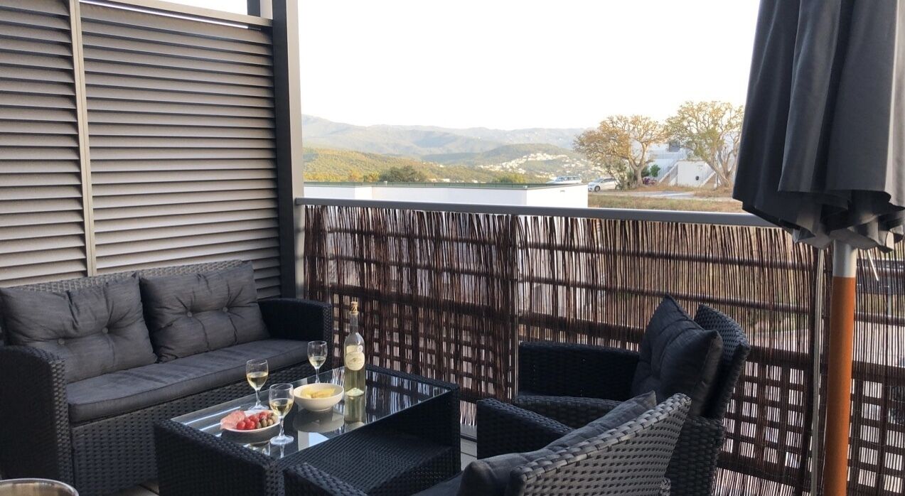 A balcony with a sofa, wicker chairs, and a glass table with a bottle and bowl of fruit.