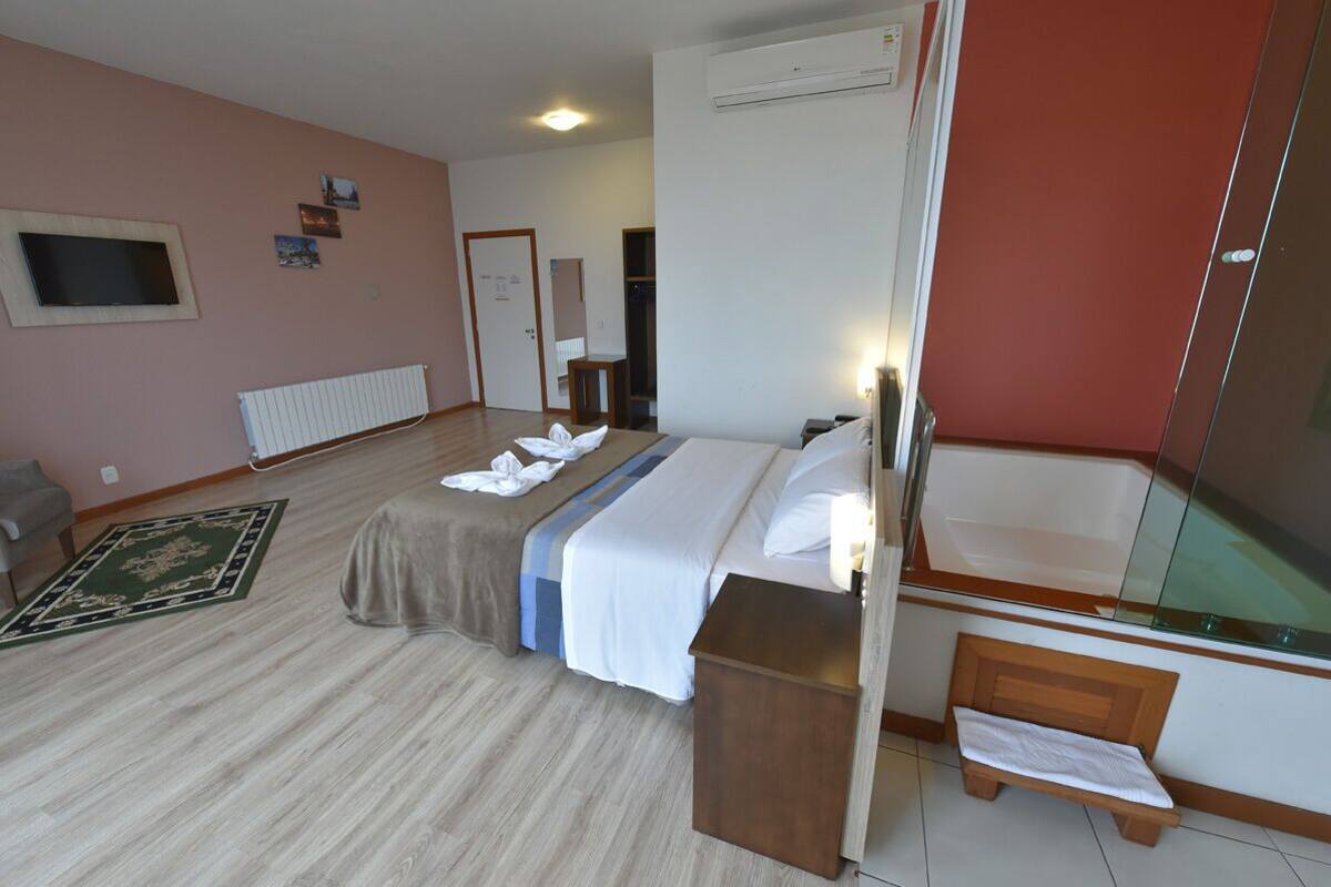 A hotel room with a bed, a small bedside table, a mirror, a radiator, a TV, and a door leading to another room.