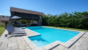 Une piscine rectangulaire, bordée d’une terrasse carrelée, entourée d’une pelouse et d’un bâtiment en bois doté d’un toit en tuiles.