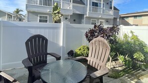 A patio with two Adirondack chairs and a glass table, a white fence, and a two-story house with a balcony in the background. - Mandalay Shores Resort (Oxnard)