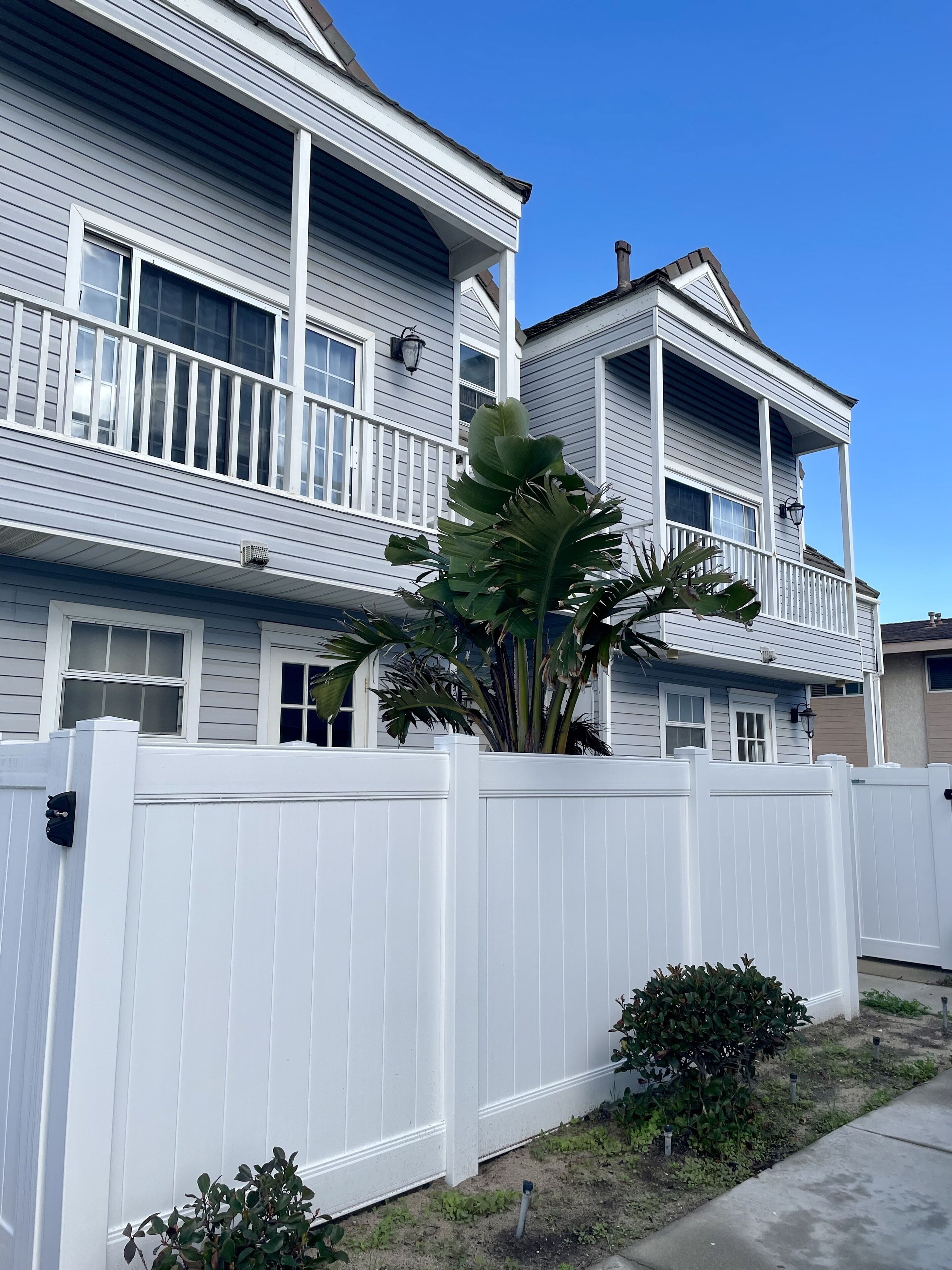 A two-story house with a white picket fence, a balcony, and a large plant in the foreground.