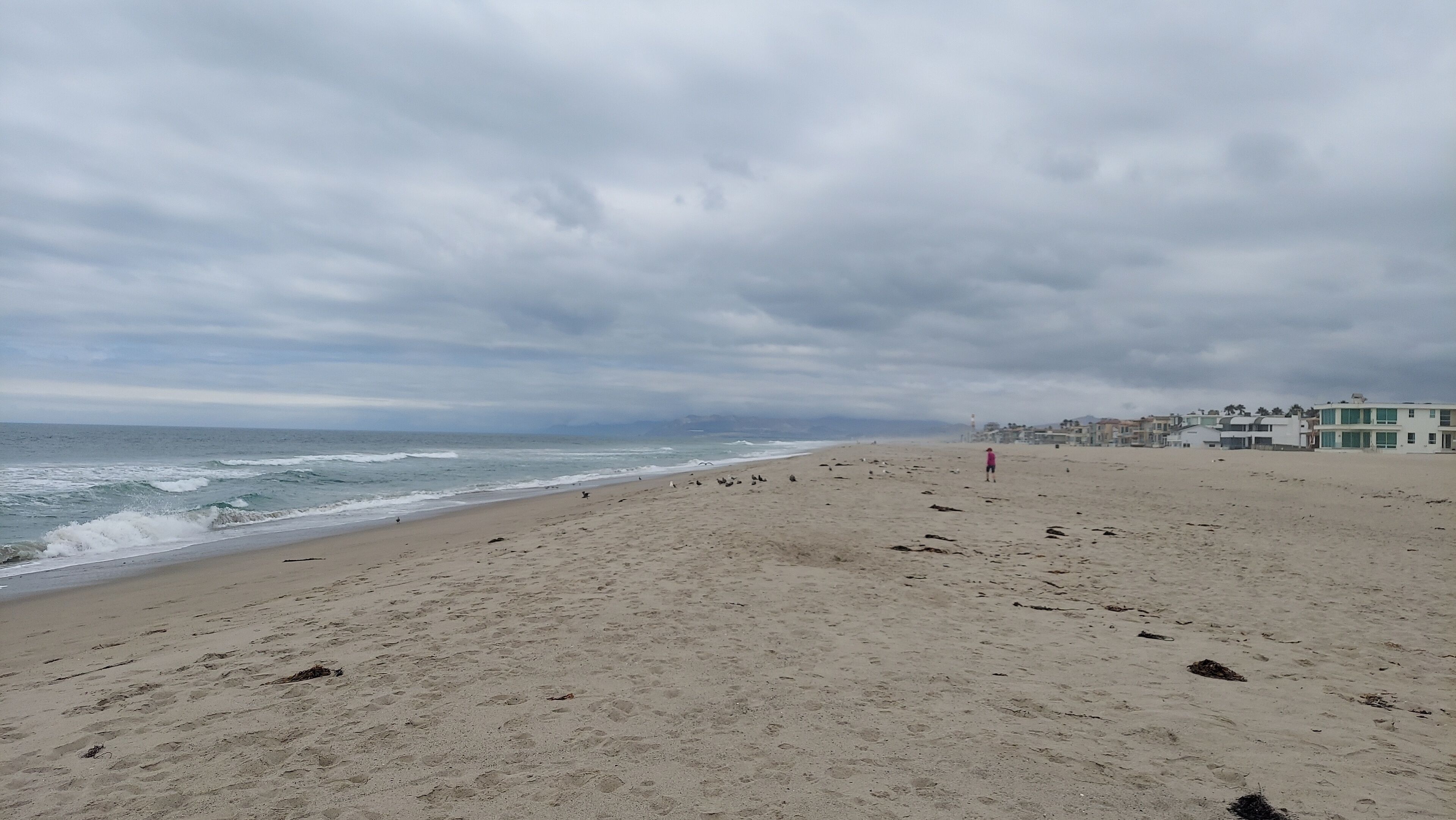 Uma extensa praia de areia com ondas do oceano, céu nublado e edifícios ao fundo.