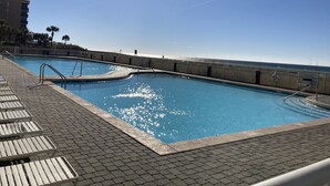 A swimming pool with a wooden deck and lounge chairs, surrounded by a wooden fence with a lifebuoy, and a clear view of the ocean. - Updated Coastal Condo • Steps to Beach • Pool • King + Bunks • Ground Floor (Fort Walton Beach)