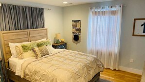 Quarto com cama de madeira, mesa de cabeceira com abajur, um objeto pendurado na parede e janela com cortinas.