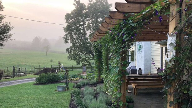 Uma área de estar rústica ao ar livre, com pérgola de madeira, caminho de pedra e vista para um campo nevoento.