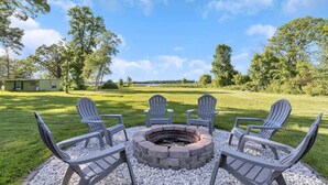 A circular fire pit with four Adirondack chairs arranged around it, set on a gravel surface with a grassy field and trees in the background. - Lake Front Home with Pool and Stunning Views (Annandale)