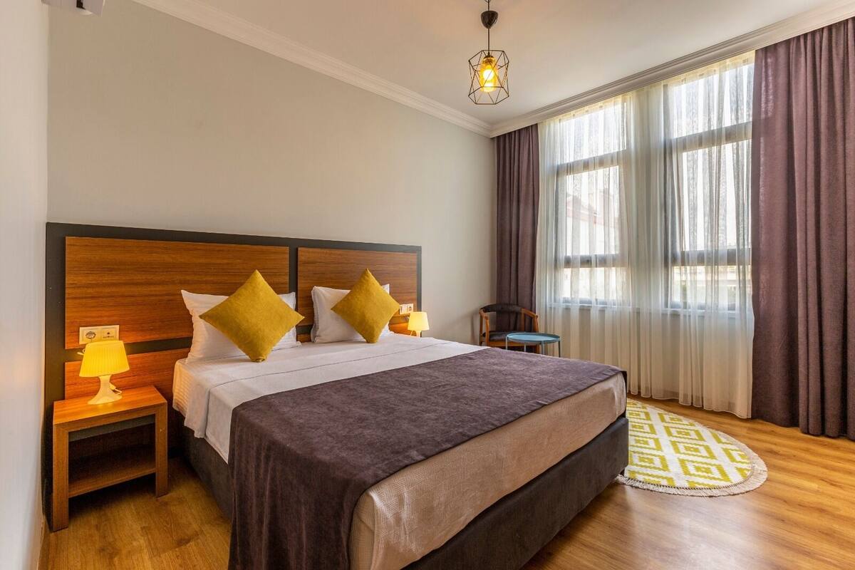 Ein Hotelzimmer mit einem großen Bett, zwei Nachttischen mit Lampen, einem Sessel, einem kleinen Tisch und einem Fenster mit Vorhängen.