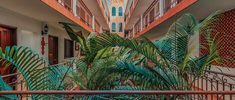 Un patio con una escalera central, rodeado de exuberante vegetación tropical y vistas a un edificio con ventanas arqueadas.