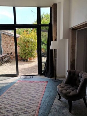 Ein Zimmer mit einer großen Schiebeglas-Tür, die auf eine Außenpatio mit Steinmauer und einem Tisch mit Stühlen führt. Auf dem Boden liegt ein Teppich mit Muster, und es steht ein Sessel mit einer Lampe darin.