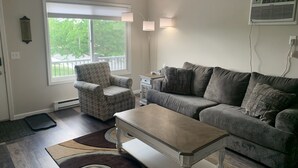 Un salón con un sofá gris, un sillón estampado, una mesa de centro de madera y un ventanal con vistas a zonas verdes.